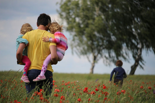 Man holding 2 kids in a field, a boy walks in front of him in a distance headed towards the trees.